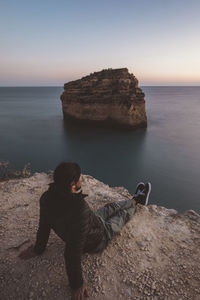 High angle view of man looking at sea while sitting on rock formation against clear sky during sunset