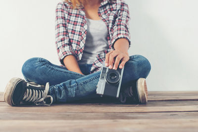 Low section of woman with camera sitting against wall on wooden floor