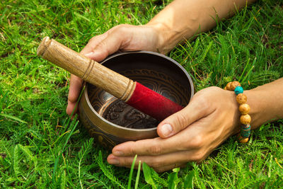 The hands of a young woman on tibetian singing bowl with wooden stick on the grass, top view.