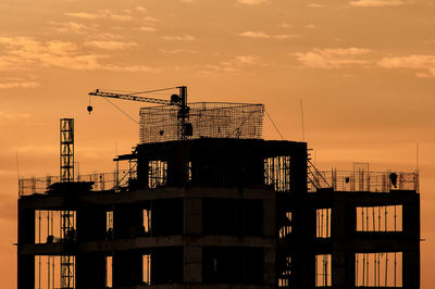 Low angle view of silhouette crane by building against sky during sunset