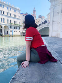 Woman sitting over river by buildings in city