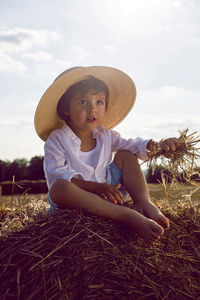 Baby boy in straw hat and blue pants sitting on a haystack in a field in autumn