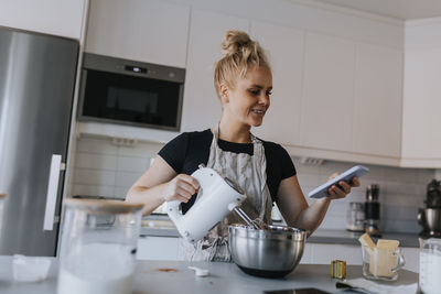 Young woman using hand mixer in kitchen and checking cell phone