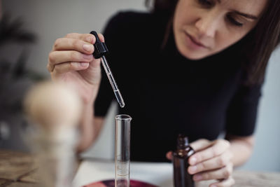 Female owner using pipette for mixing liquid from bottle while sitting at table in perfume workshop