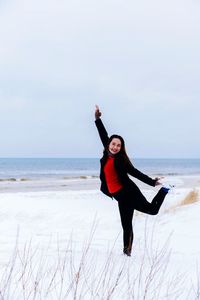 Portrait of woman standing on one leg at beach against sky during winter