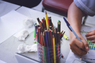 Cropped hand of boy writing on book by colorful pencils in container