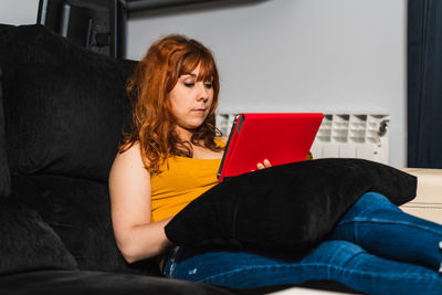 Caucasian woman sitting on the couch with her red tablet at home, dressed in yellow shirt and jeans