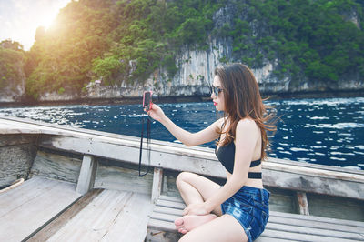 Side view of young woman taking selfie with camera while sitting in boat on river