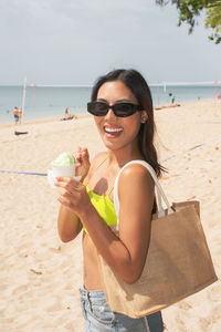 Portrait of young woman holding sunglasses at beach against sky