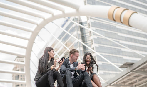 Smiling business colleagues using smart phone while sitting against buildings in city