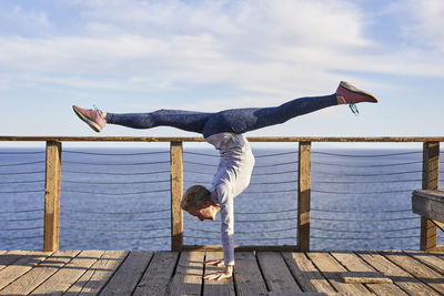 Full length senior woman doing handstand while exercising on floorboard by sea against sky