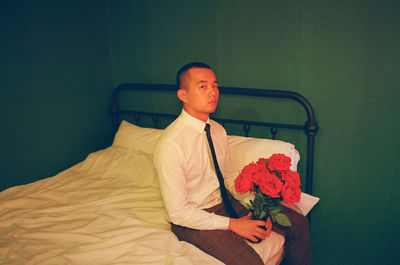 Portrait of man holding rose bouquet while sitting on bed at home