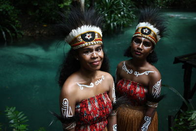 Native papua women with traditional crown headwear  standing against trees at a riverbank.