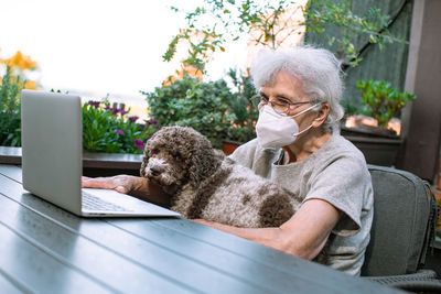 Old woman wearing face mask using laptop on terrace