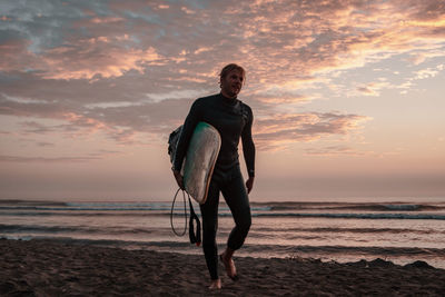 Full length of man carrying surfboard at beach against sky during sunset