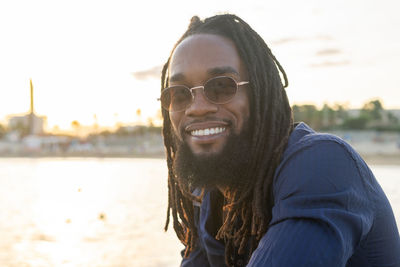 Side view of happy african american man with dreadlocks and sunglasses sitting on seashore and looking at camera against cloudless sky in summer