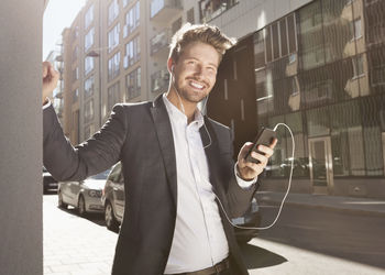 Happy young businessman listening music through mobile phone on street