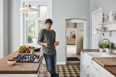 Man using smartphone and holding bell pepper in kitchen