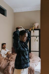 Non-binary person tying hair while standing in bedroom at home