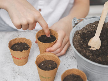 The hand of a teenage girl pushes a cucumber seed into the soil in a cardboard glass with her finger