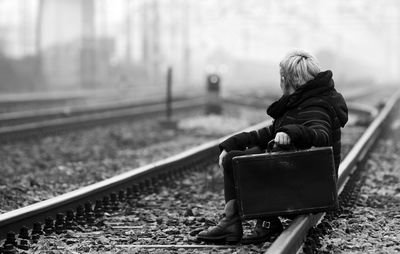 Side view of woman with suitcase sitting on railroad track