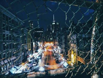 Vehicles parked on illuminated street amidst buildings seen through metallic fence at night