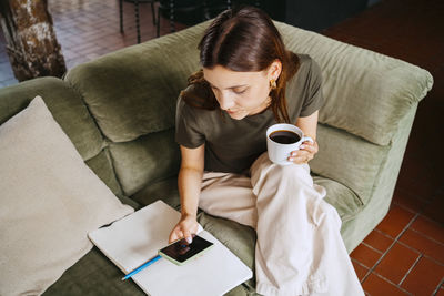 Young female entrepreneur holding coffee cup while using smart phone on sofa in studio