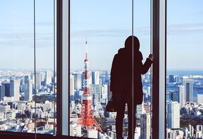 Rear view of woman standing by window against tokyo tower in city