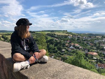 Full length of teenage girl sitting on retaining wall against sky