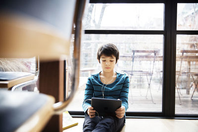 Boy using digital tablet while sitting by window at home