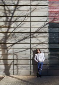 Young disabled man with hands in pockets standing against wall with tree shadow