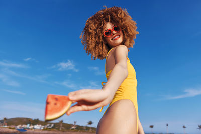 Portrait of young woman with arms raised standing against blue sky
