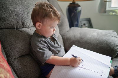 Young boy drawing doodles in a notebook while sitting on a couch at home