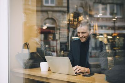 Smiling businessman using laptop in cafe seen through glass window