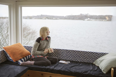 Thoughtful woman having coffee while looking at lake through window during winter