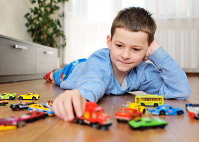 Preschool boy in a blue sweater is lying on the wooden floor playing with toy cars 