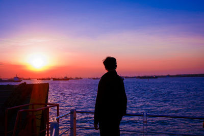 Silhouette man looking at sea while standing by railing against sky during sunset