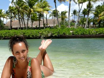 Full length portrait of happy woman lying on shore in sea against palm trees