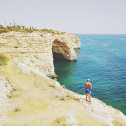 Rear view of man standing on cliff by sea against clear sky