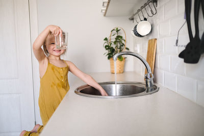 European child of 4 years old washes glass in sink on his own, bright kitchen in real interior, 