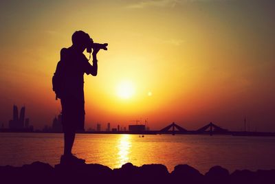 Silhouette man photographing by river against sky during sunset