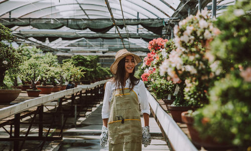 Smiling woman wearing hat standing amidst potted plants in greenhouse