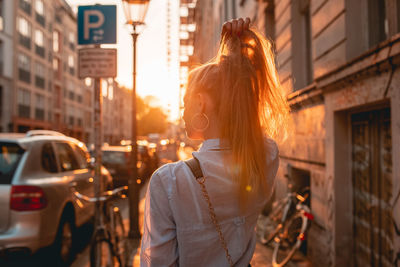 Rear view of young woman with hand in hair while standing on sidewalk during sunset