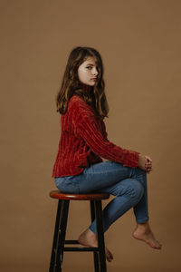 Portrait of serious girl sitting on stool against brown background