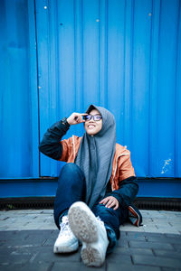 Full length of young woman sitting against blue wall