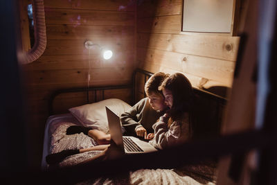 Brother and sister using laptop while sitting in bedroom against wall at home