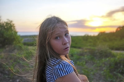 Portrait of teenage girl with long blond hair on field against sky during sunset
