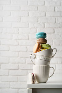 Stack of white coffee cups filled with macaroons on the edge of the table, white brick wall backgro