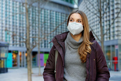 Medium shot of beautiful woman wearing protective face mask walking in city street in winter clothes