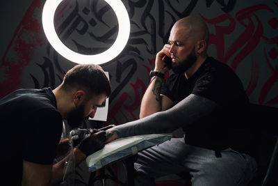 Serious male tattoo master using tattoo machine while making tattoo on hand of guy with phone in light tattoo studio
